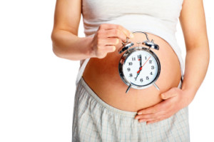 Mid section of a young woman holding an alarm clock against her pregnant belly on white